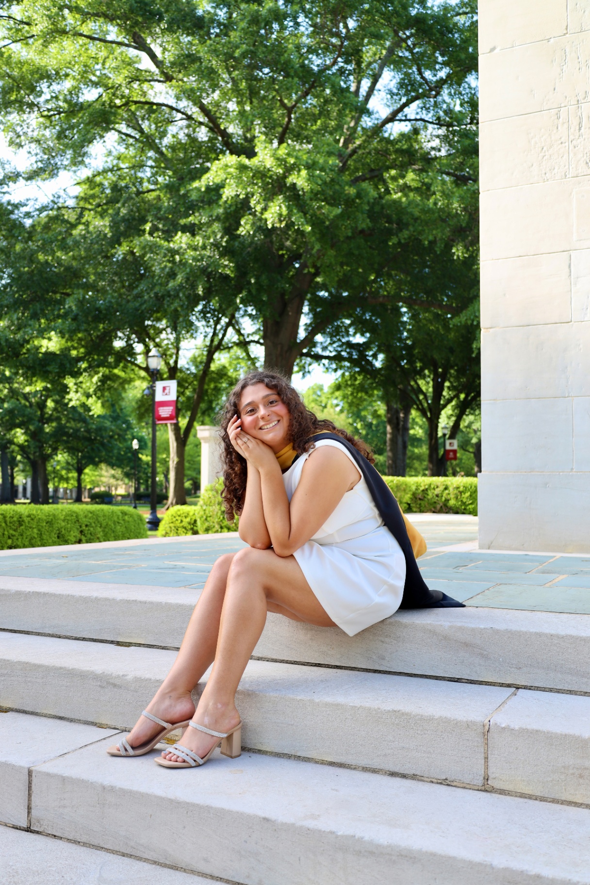 Graduate seated on campus steps in a white dress and honors stole