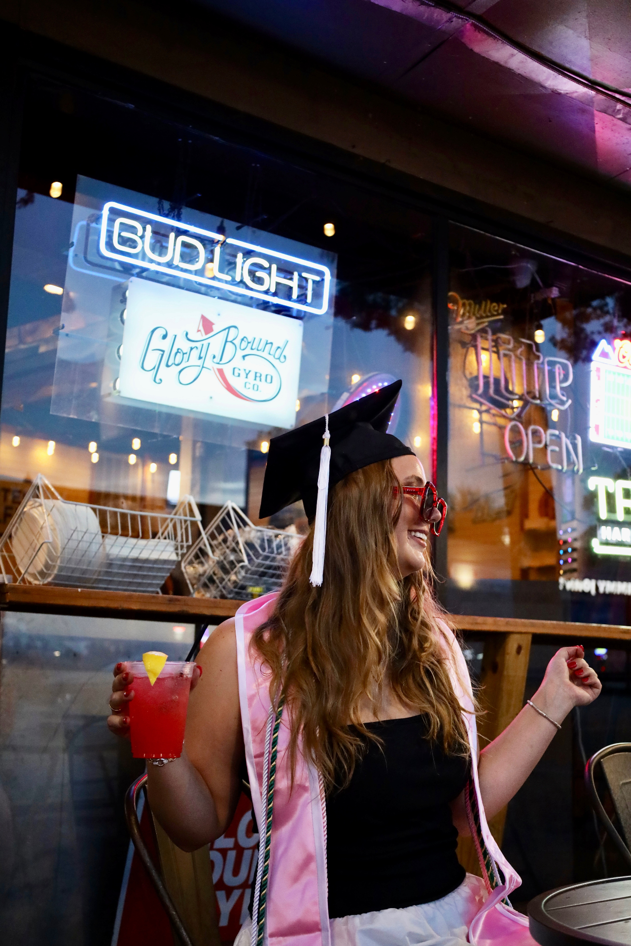 Graduate in cap and red sunglasses with a cocktail beneath neon signs