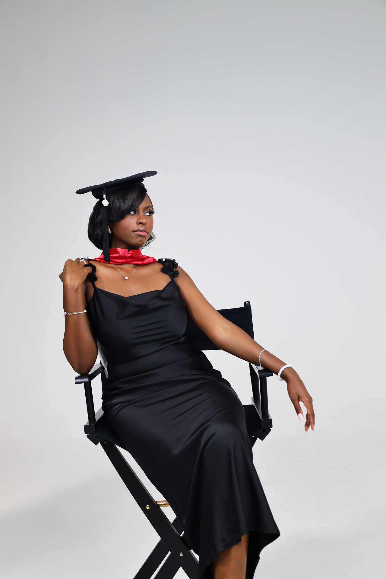 Graduate studio portrait in black satin dress and cap seated in a director chair