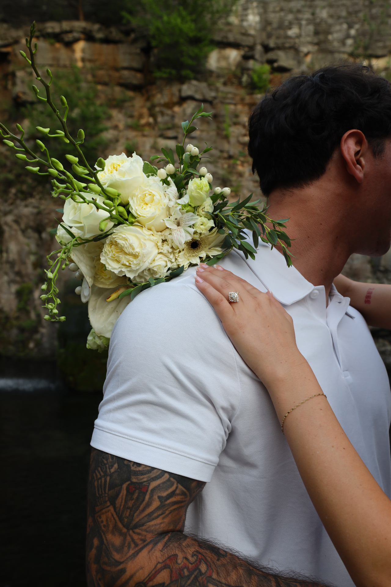 Bride's hand with engagement ring resting on groom's shoulder beside a white bouquet