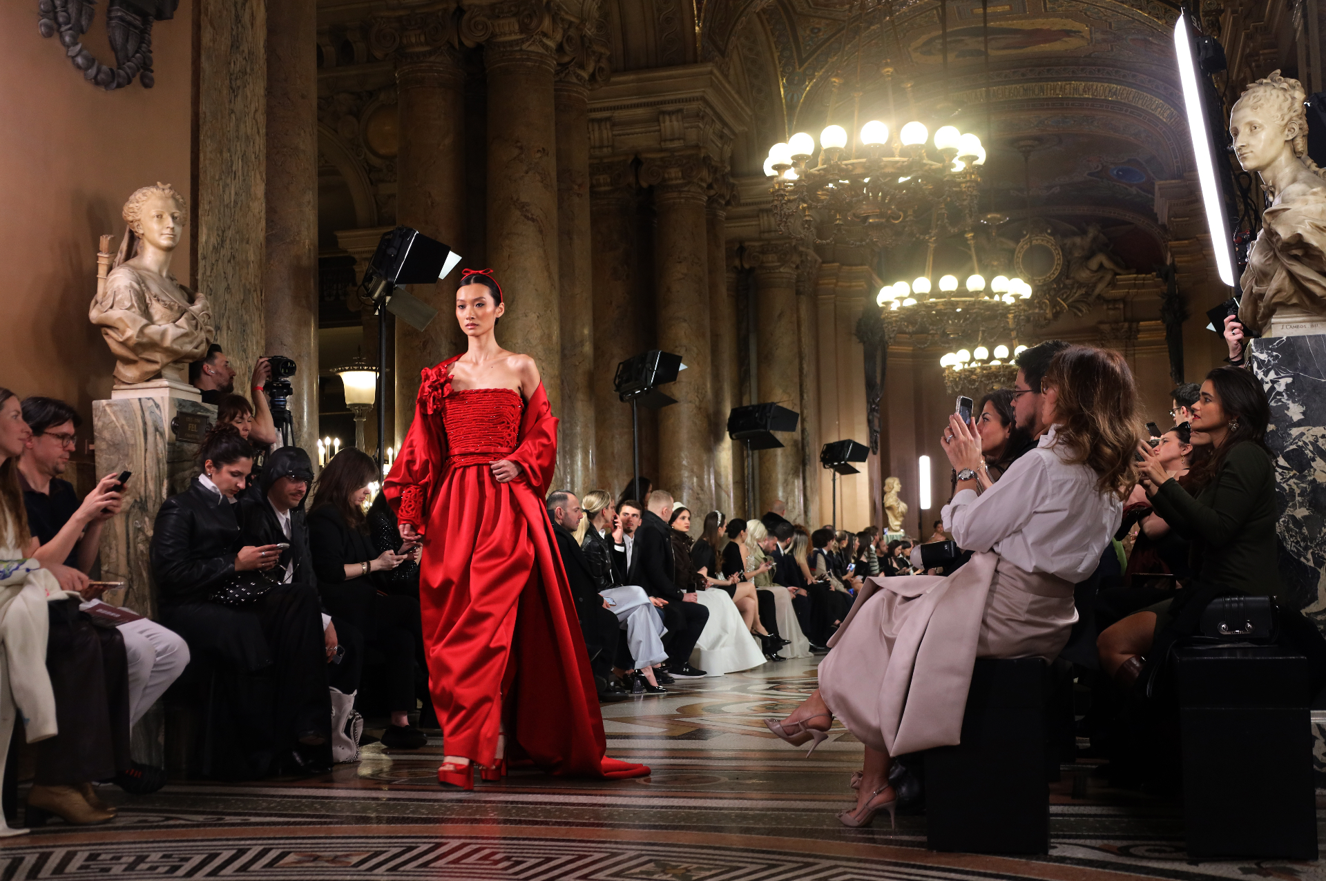 Wide runway view with model in red gown beneath chandeliers